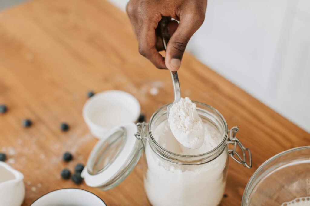 A person scooping flour from a glass jar on a wooden table surrounded by blueberries.